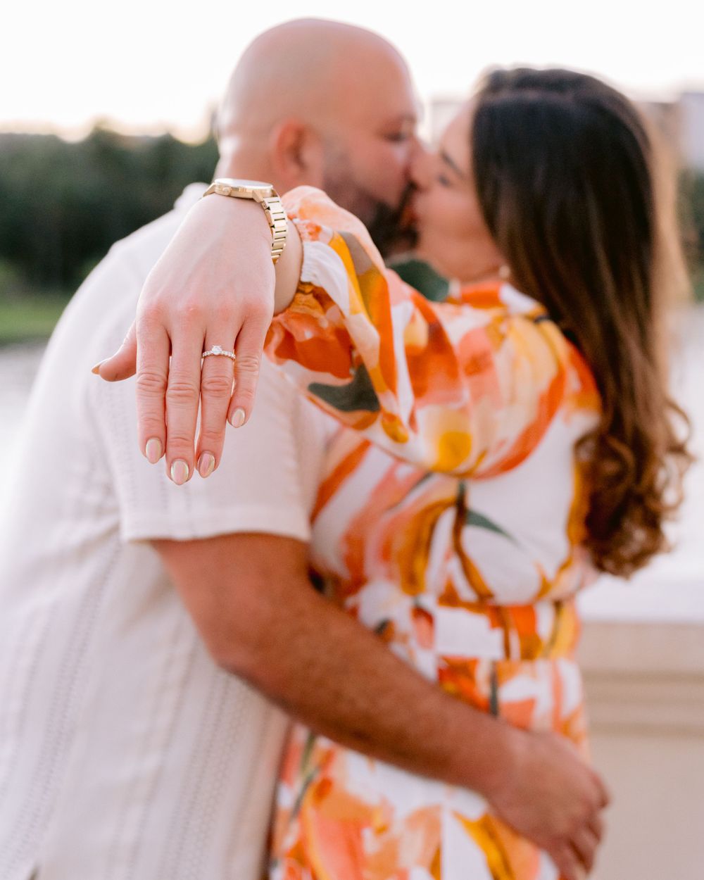 A couple shares a romantic moment while showing off an engagement ring in orange floral attire outdoors at sunset.