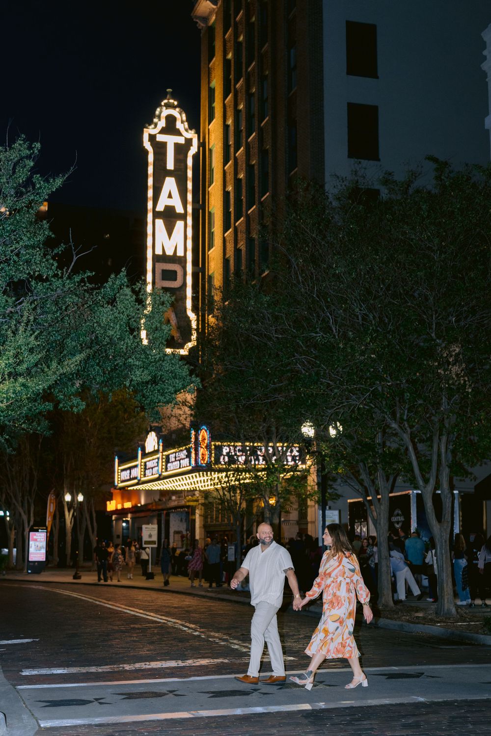 A couple crosses the street at night in front of the illuminated Tampa Theatre's historic marquee and neon sign.