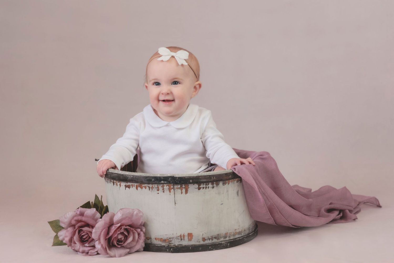 A smiling baby wearing white sits in a vintage prop with dusty pink roses. Tiny moments photography, Hampshire baby photographer
