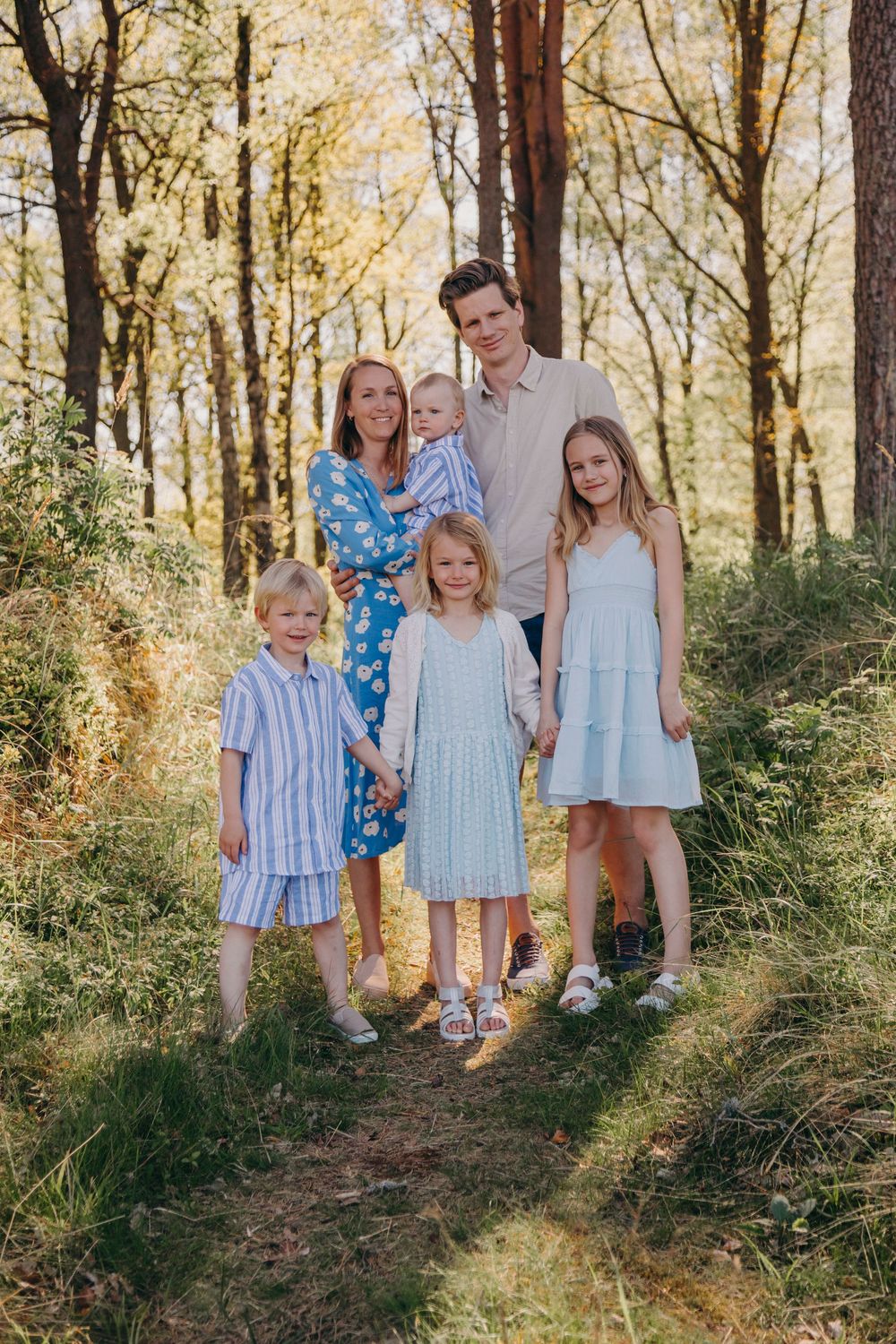 A family poses together on a forest path during a golden autumn afternoon wearing coordinated blue and white outfits.