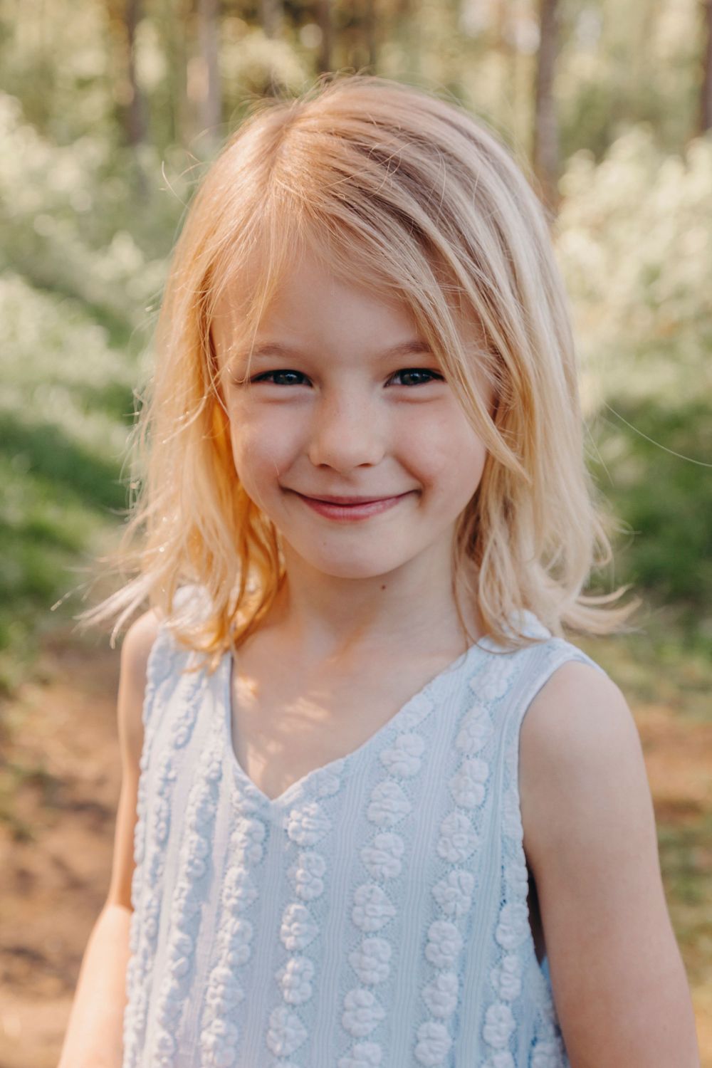 Child with blonde hair wearing a light blue sleeveless dress smiles outdoors in natural lighting.