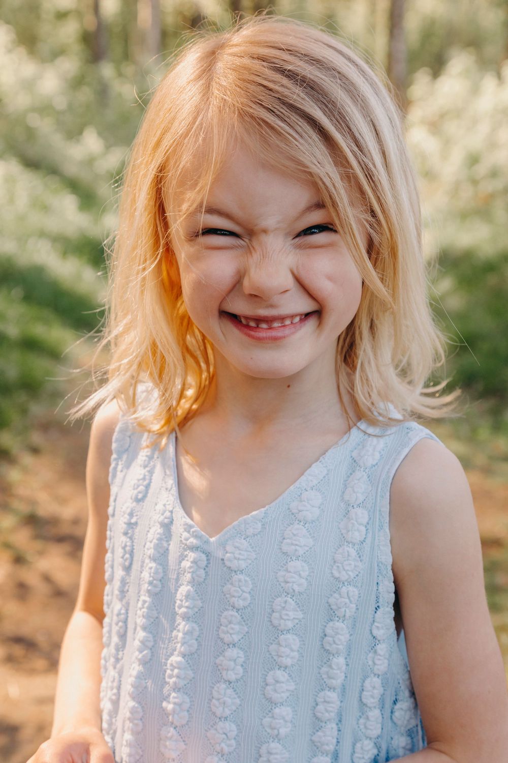 Young person in light blue lace dress smiling joyfully outdoors with natural lighting and blurred foliage background.