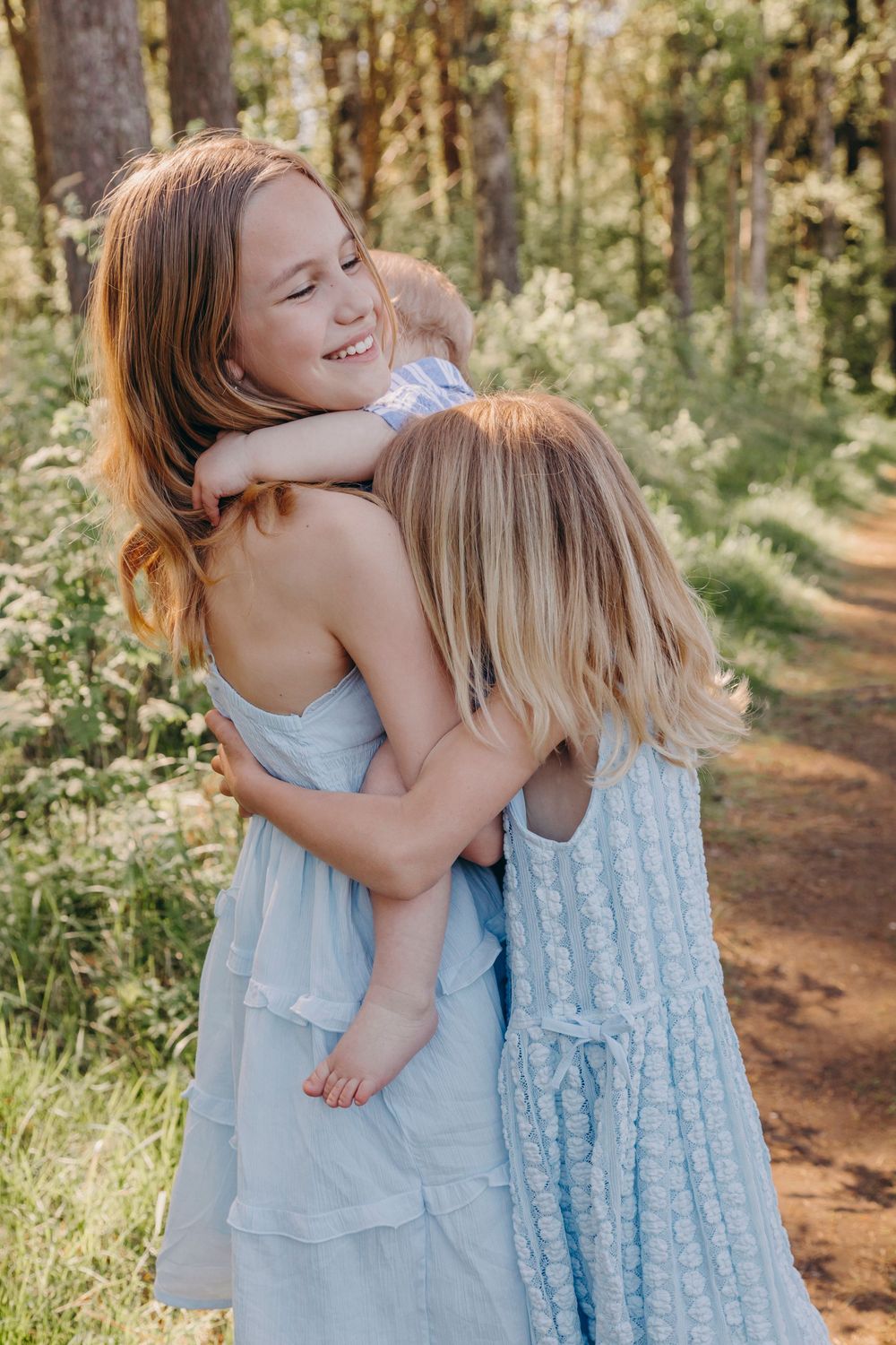 Two people in blue dresses share a warm embrace on a wooded trail during golden hour.