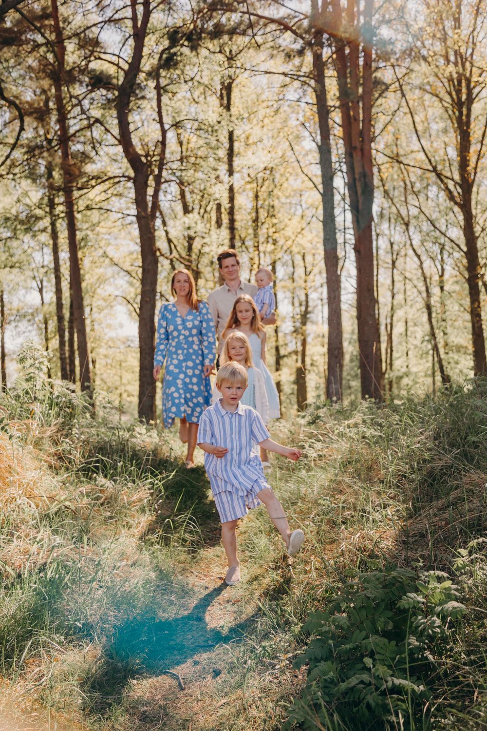 Family of four walking together on a sunlit forest trail wearing coordinated blue and white outfits.