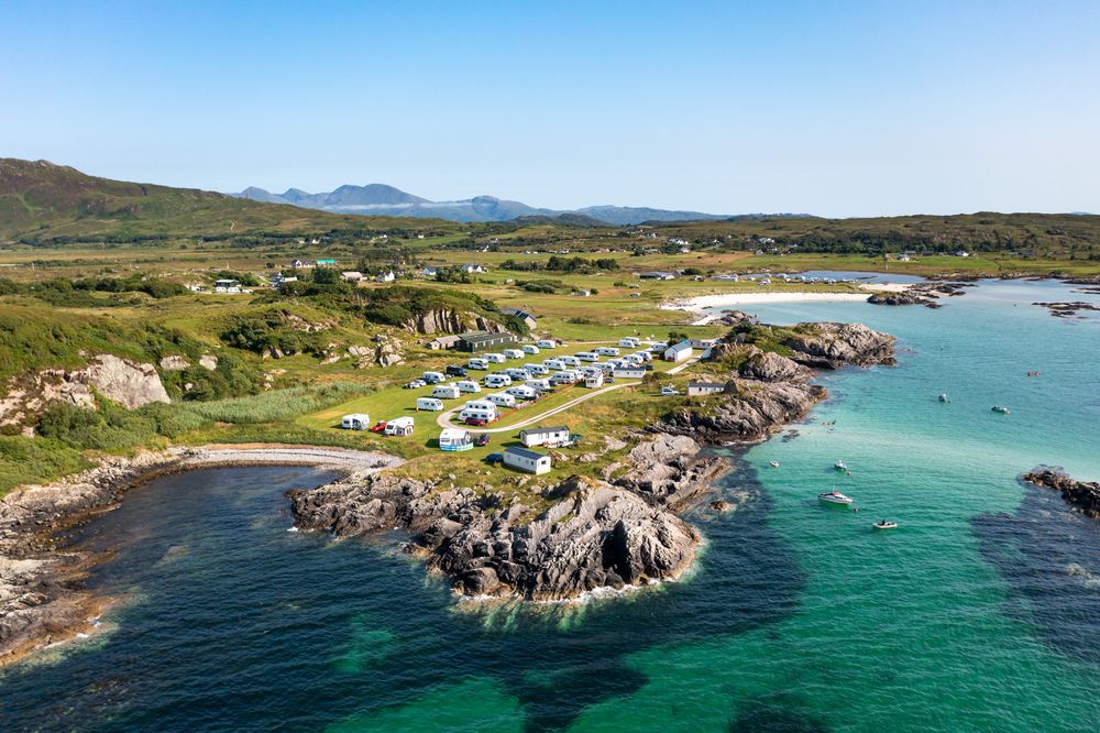 Aerial view of a rocky coastline in Scotland with turquoise waters, camping sites, and distant mountains on the horizon.