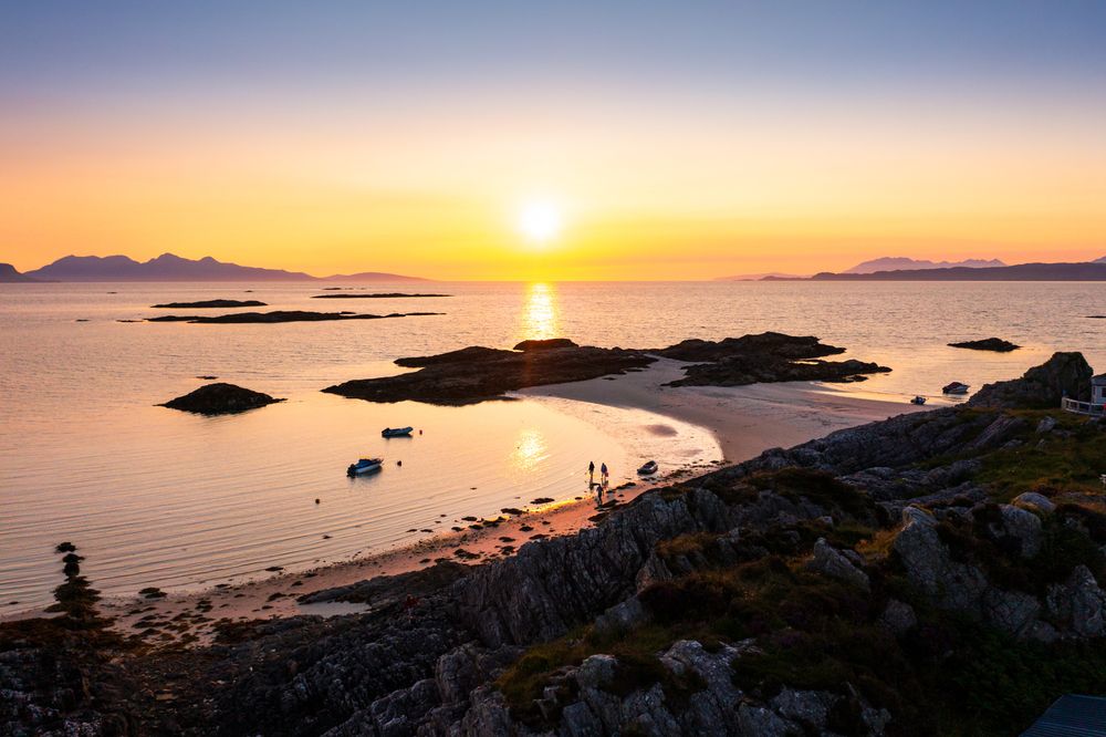 Stunning sunset over calm ocean with rocky islands silhouetted against golden sky and mountains in distance.