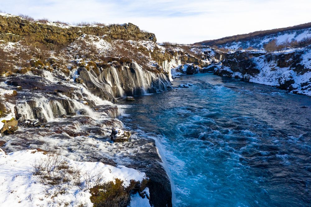 Turquoise waters flow past snow-covered rocky cliffs at Hraunfossar waterfall in Iceland during winter.