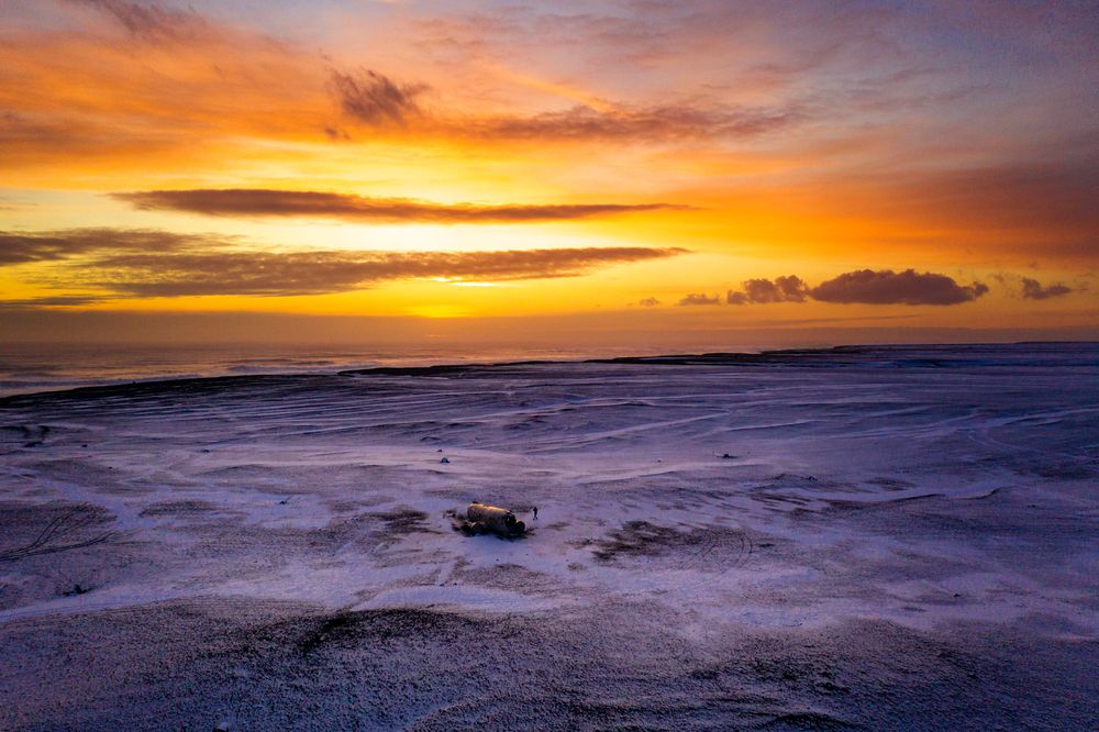 Dramatic orange and yellow sunset sky over calm ocean waves with snowy shore in winter landscape.