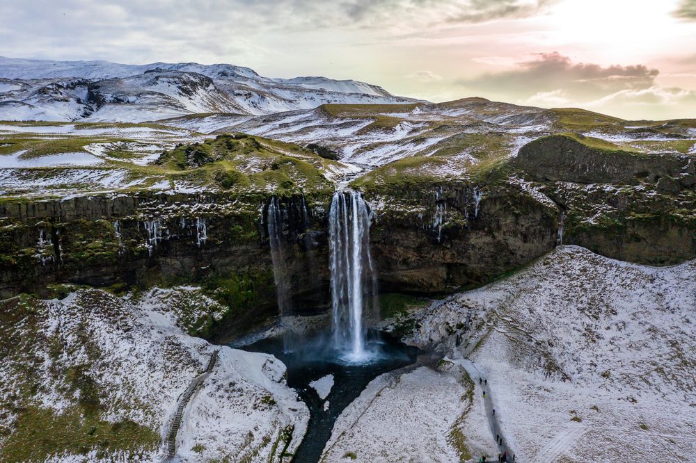 Seljalandsfoss waterfall cascades over snowy cliffs into a blue pool amid winter landscape in Iceland at sunset.