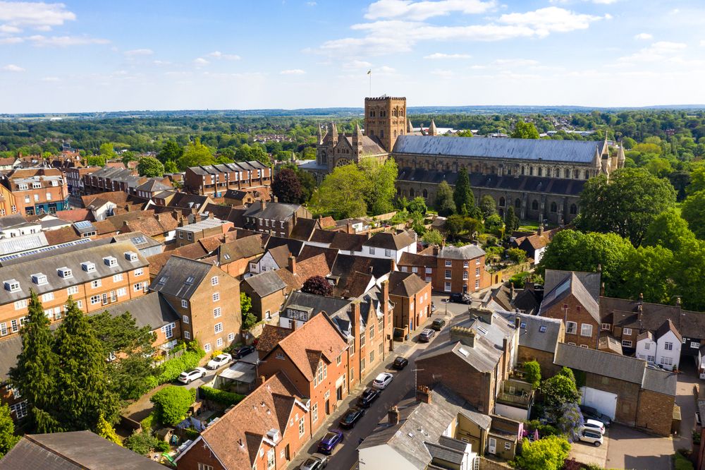 Aerial view of St Albans Cathedral and historic city rooftops surrounded by green trees on a sunny day in England.