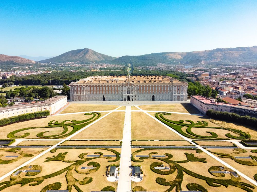 Aerial view of ornate palace gardens with symmetrical baroque patterns and grand architecture in Royal Palace of Caserta, Italy.