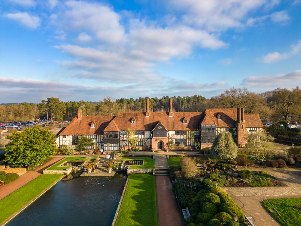 Aerial view of historic English Tudor manor house with manicured gardens and pond on a sunny day beneath blue skies.
