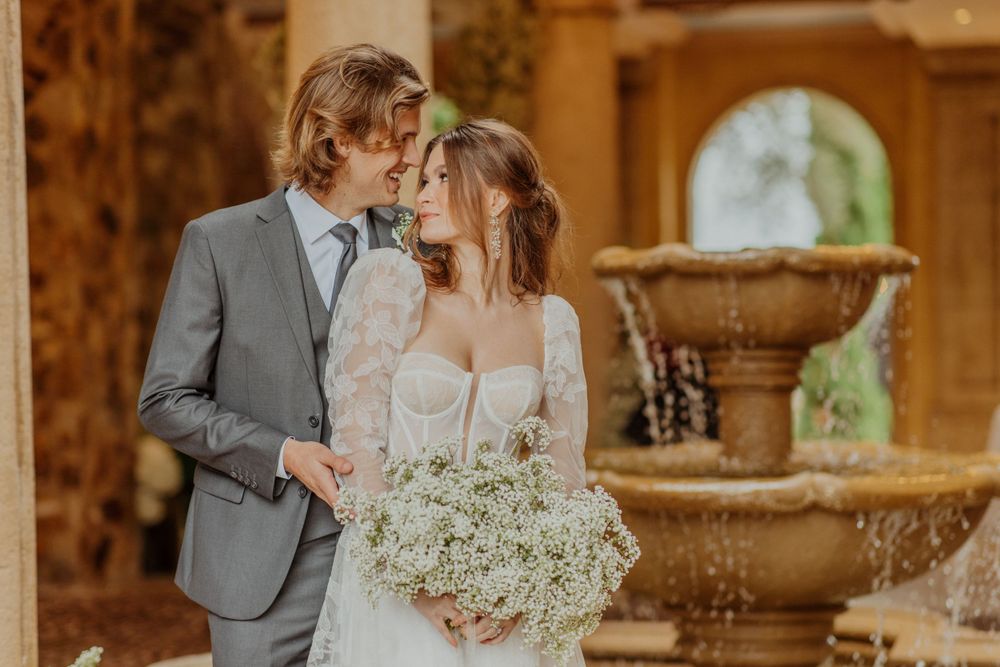 A romantic wedding couple share an intimate moment near a decorative fountain in a golden sunlit rustic setting.