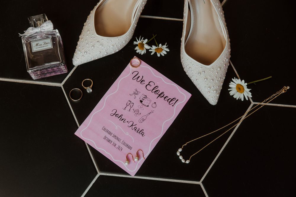 Wedding details flatlay with white sparkly heels, perfume bottle, rings, daisies and pink wedding invitation on black surface in Colorado.