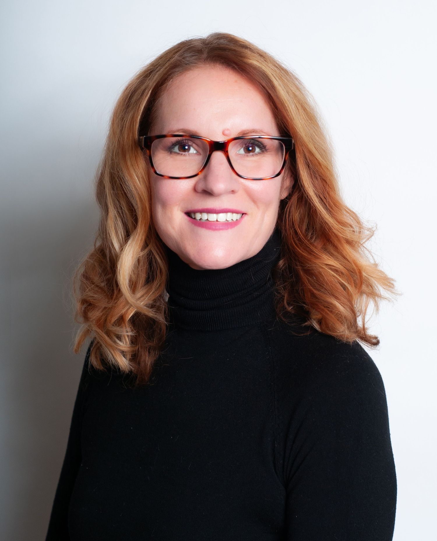 Professional headshot of someone with wavy reddish hair wearing glasses and a black turtleneck against a light background.