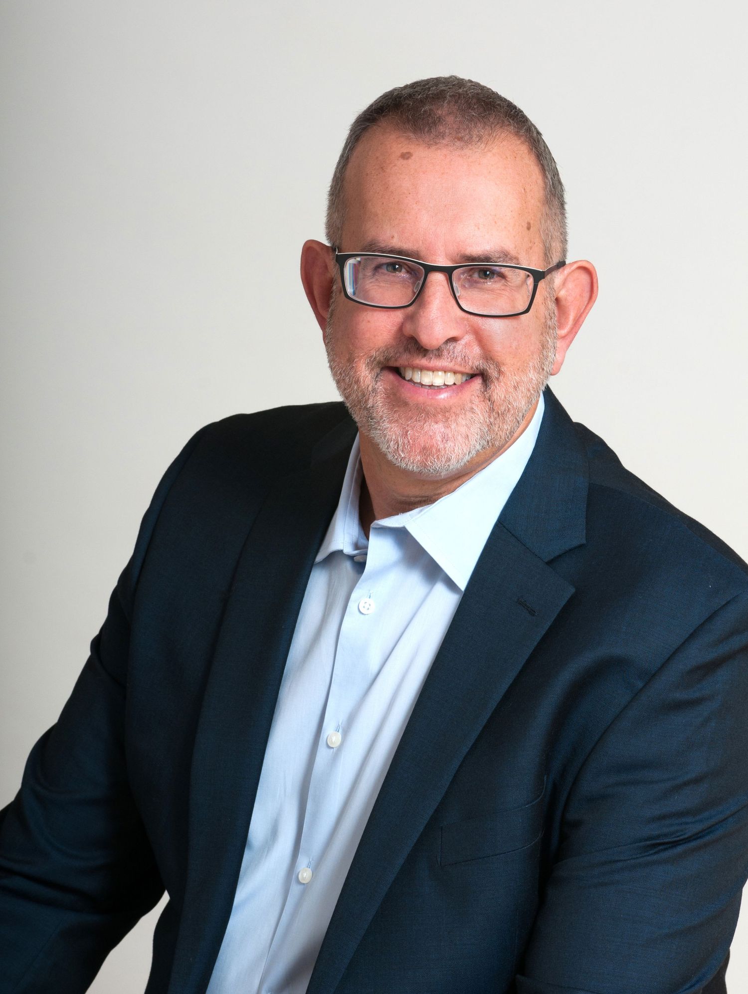 Professional headshot of a person in a navy suit and light blue dress shirt wearing glasses against a light background.