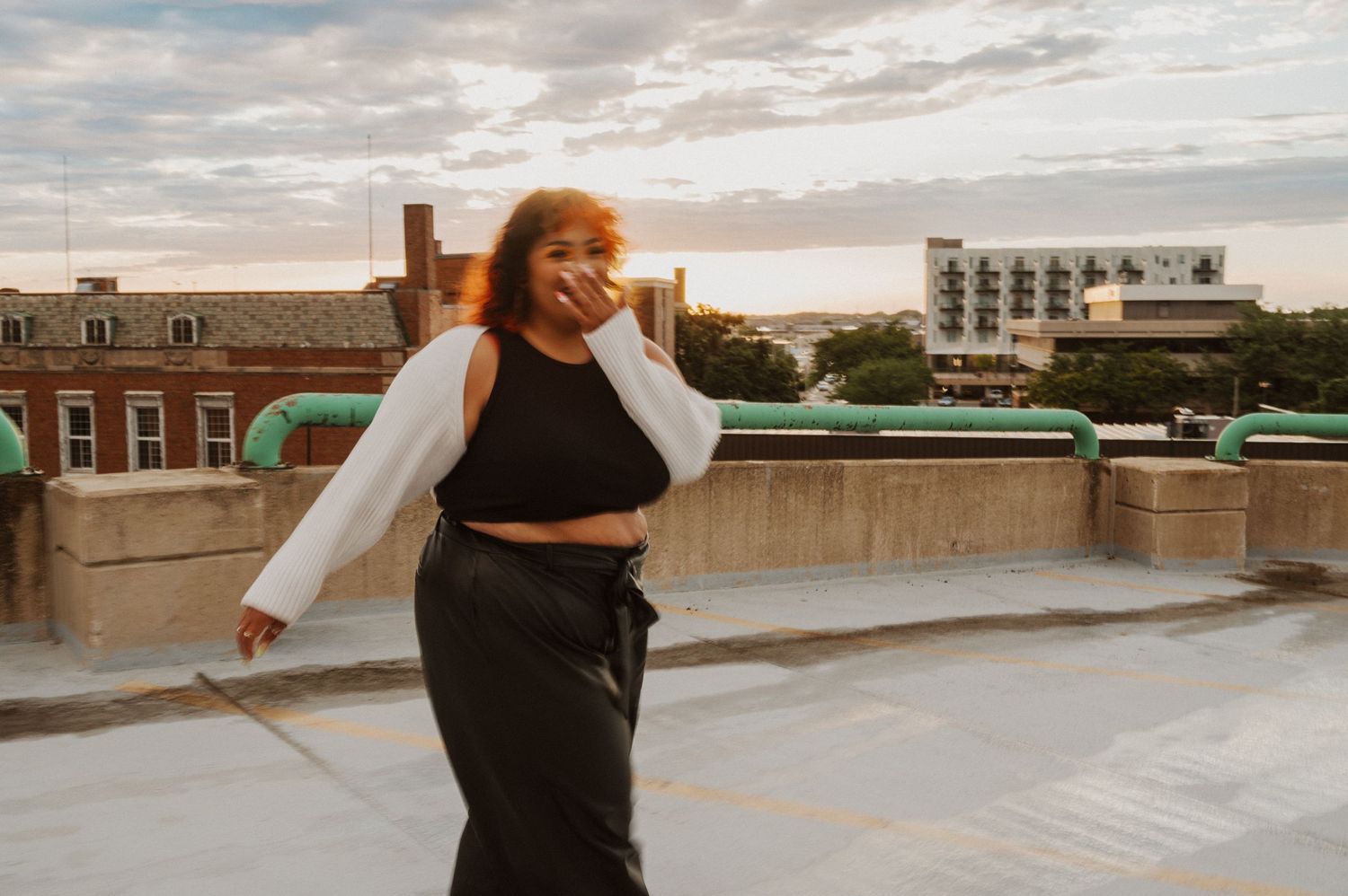 A figure in a white jacket and black crop top dances on an urban rooftop at sunset with city buildings in the background.