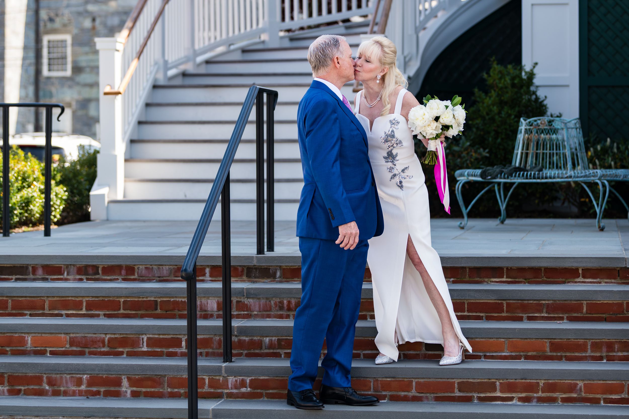 Couple embracing while walking down brick steps in elegant wedding attire on sunny day.