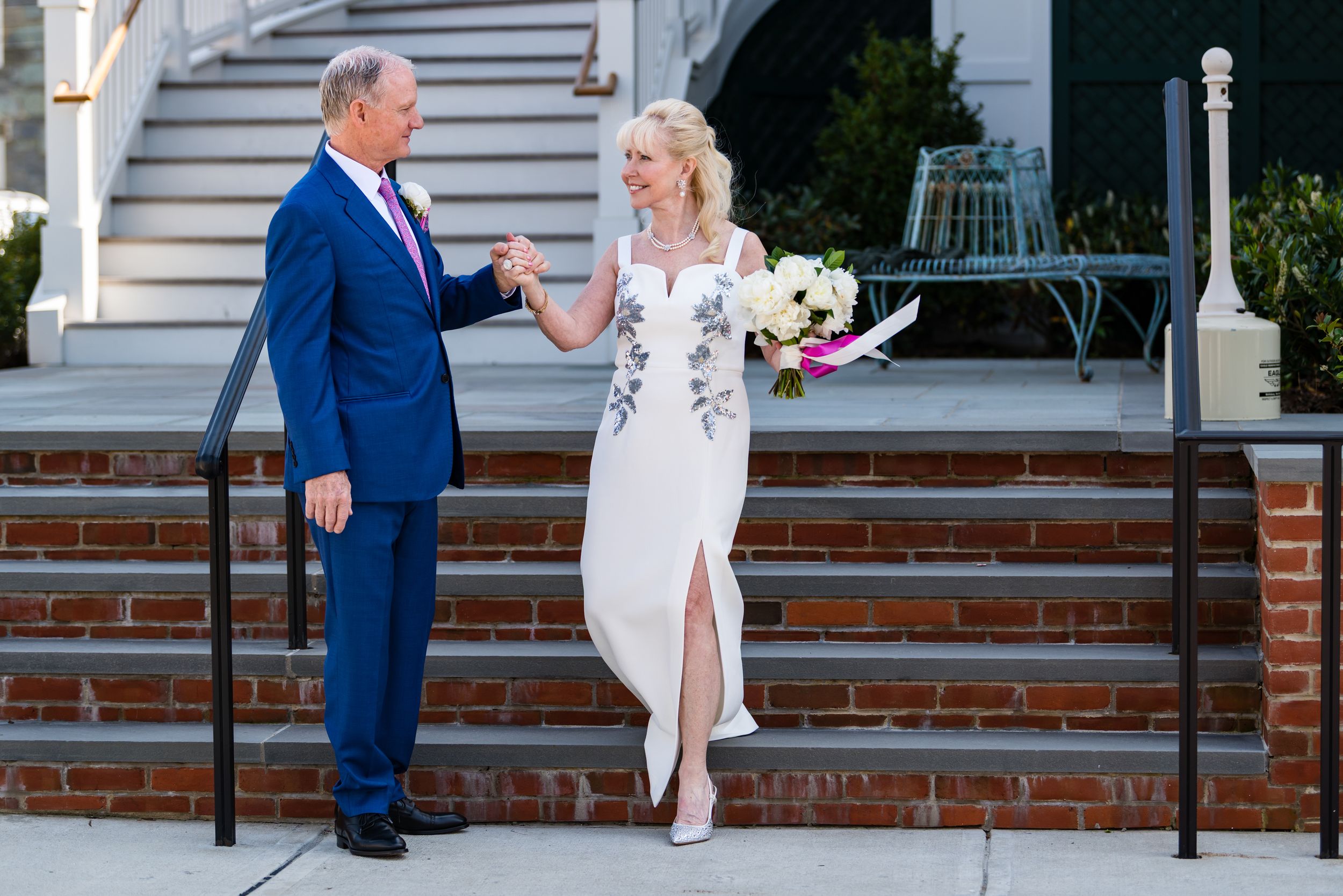 Two people in formal attire share a moment on brick steps, with one wearing a blue suit and another in a white dress.