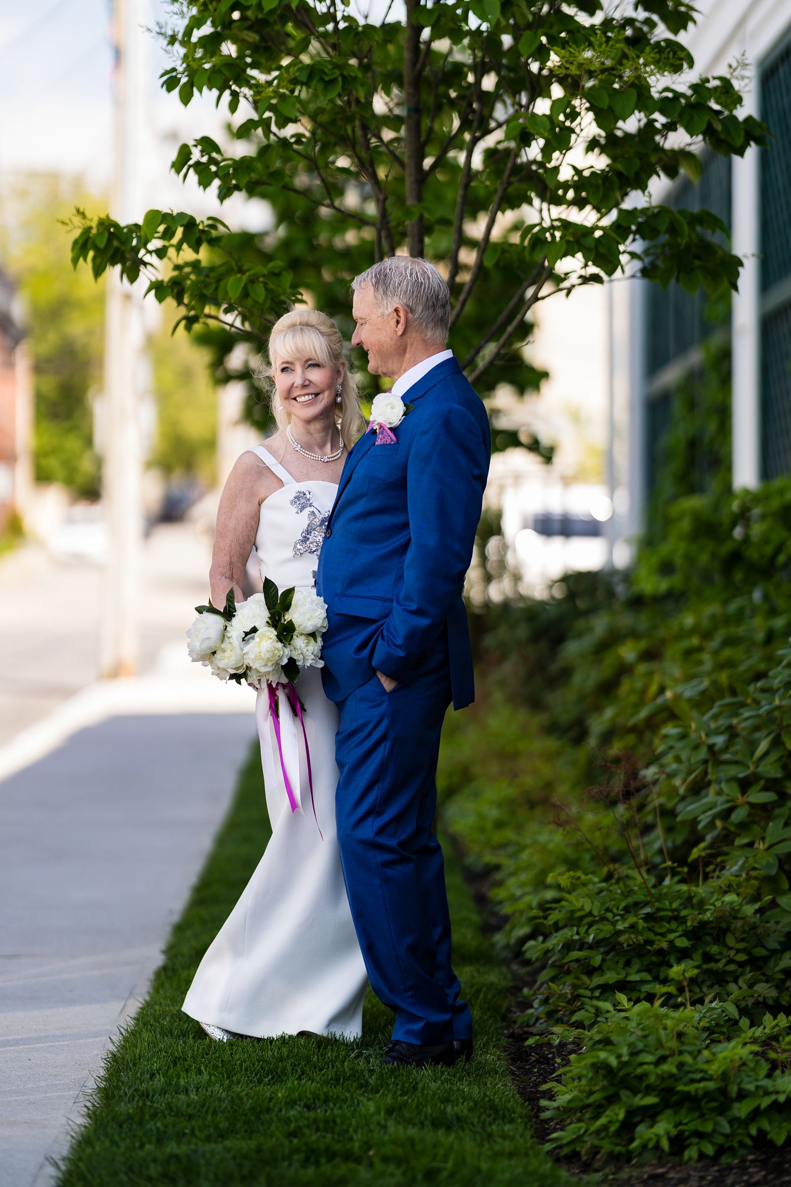 A bride and groom share a candid moment outdoors against a backdrop of trees and bushes.