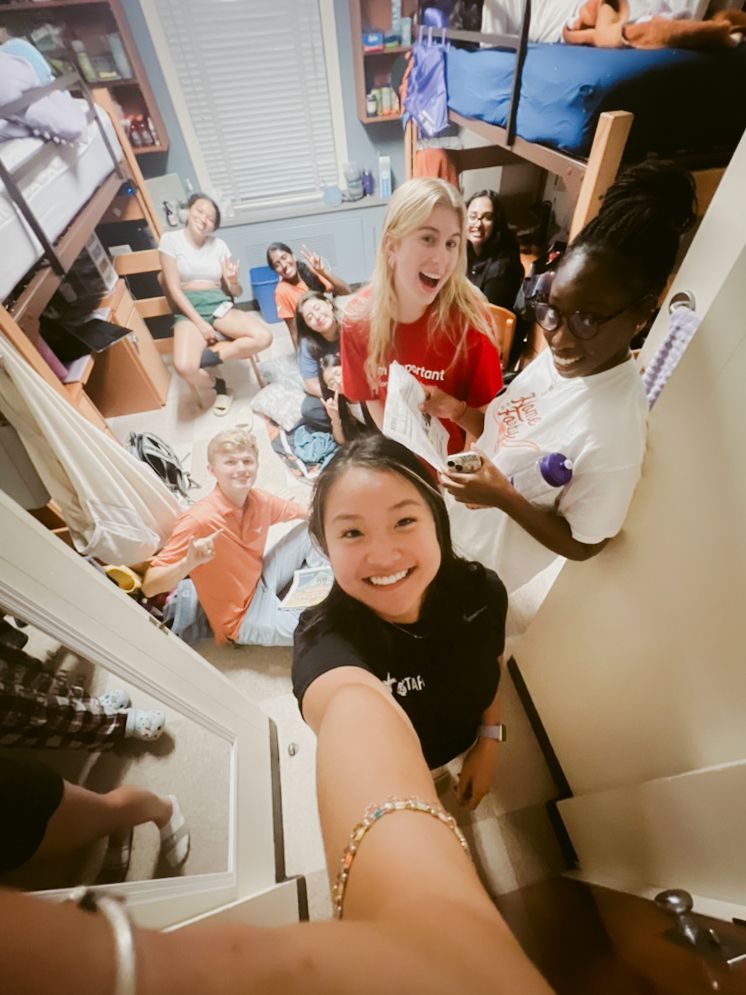 A group gathers in a cramped dorm room while someone takes a selfie-style photo from above.