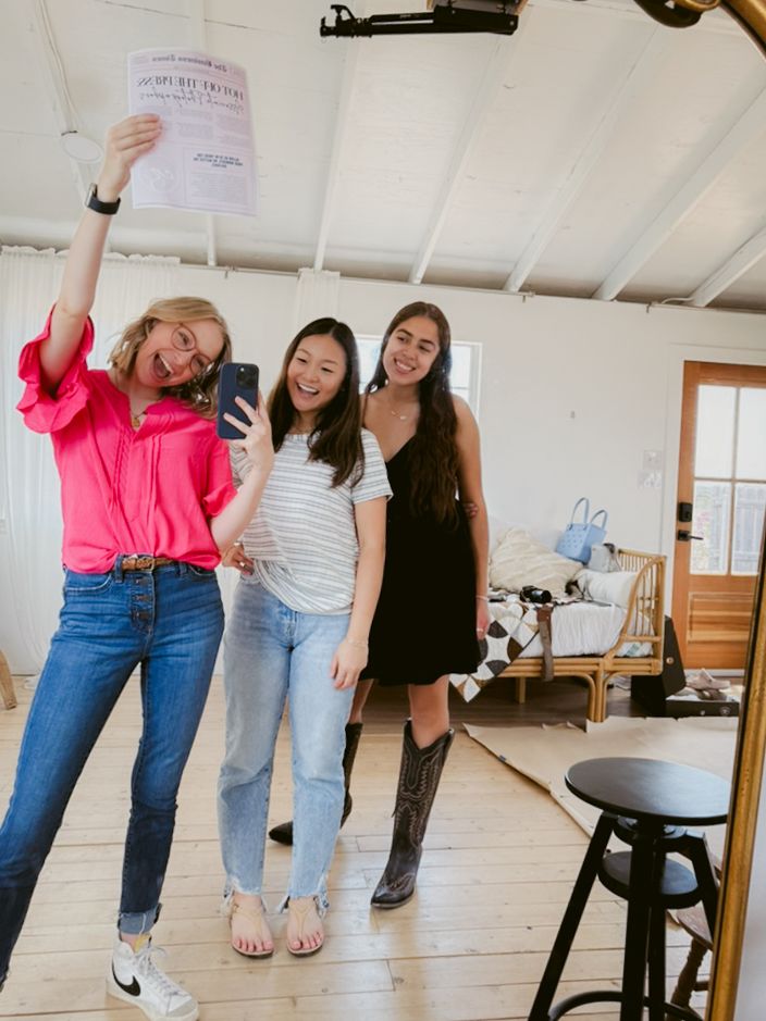 Three friends pose together in a bright indoor space with white walls and wooden floors.
