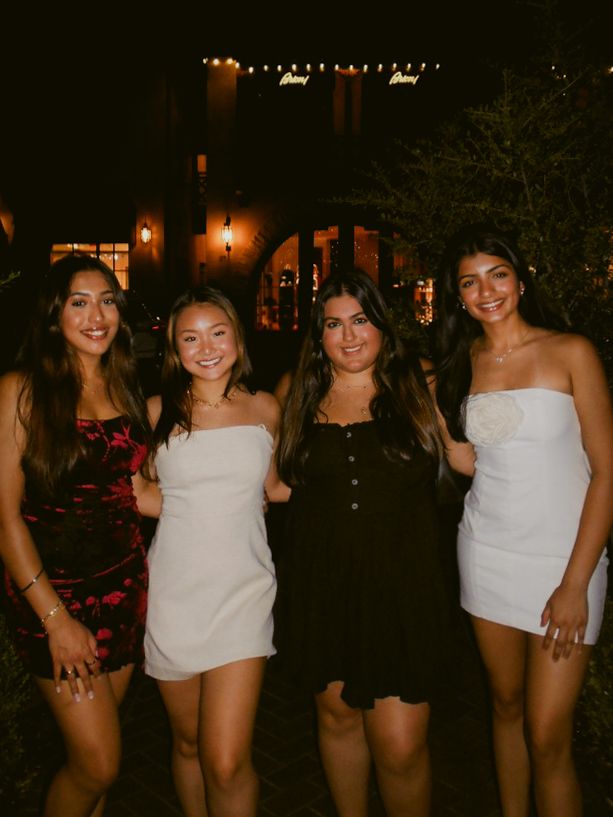 Four friends dressed up for a night out pose together under string lights at an outdoor venue.