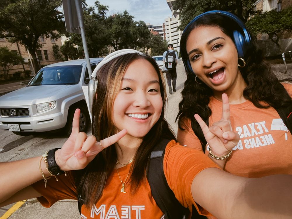 Two students in orange t-shirts taking a selfie outdoors with peace signs, one wearing blue headphones.