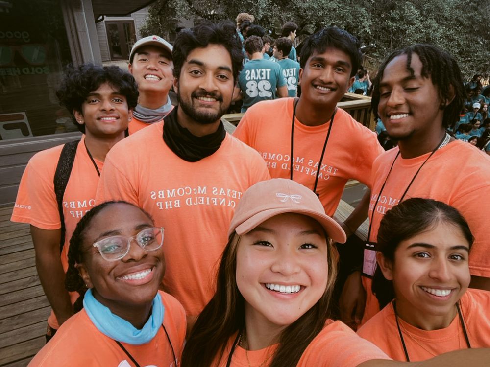 A group of volunteers wearing matching orange t-shirts smile together for a selfie photo outdoors.