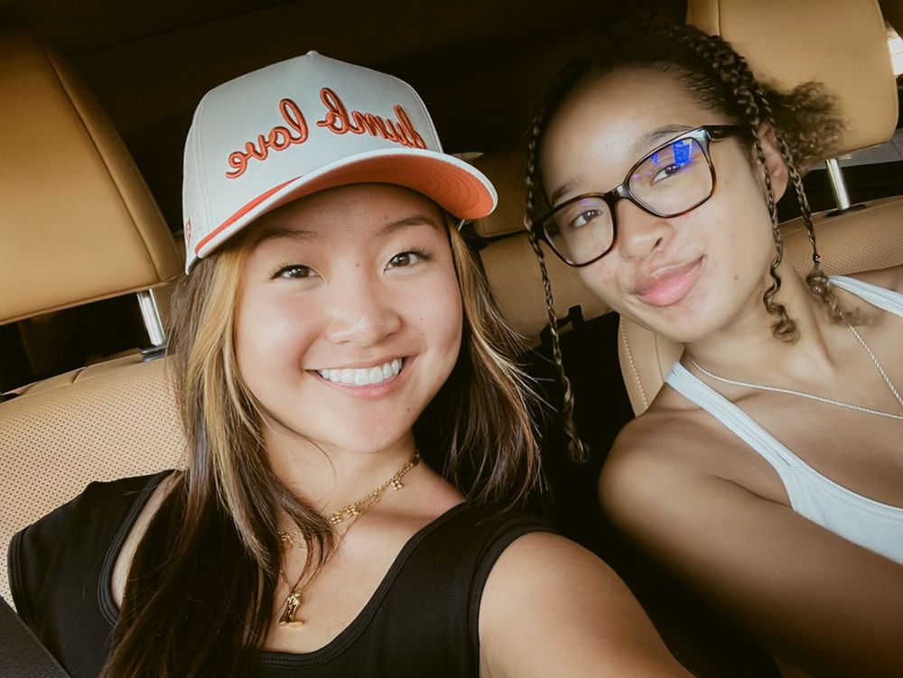 Two friends take a casual selfie together while wearing a white baseball cap and glasses.