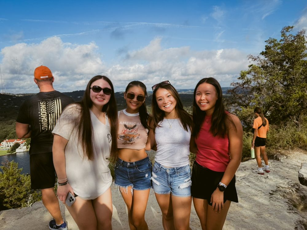 A group of friends pose together on a rocky overlook during a sunny day hike with blue skies and trees in the background.