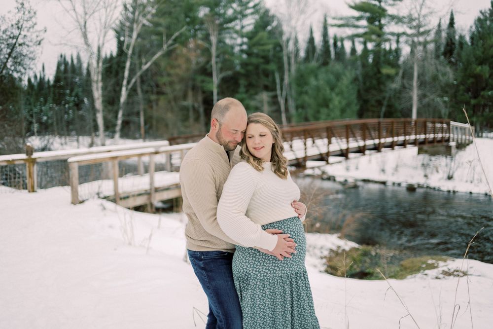 A winter maternity photoshoot by a snow-covered bridge over flowing water.