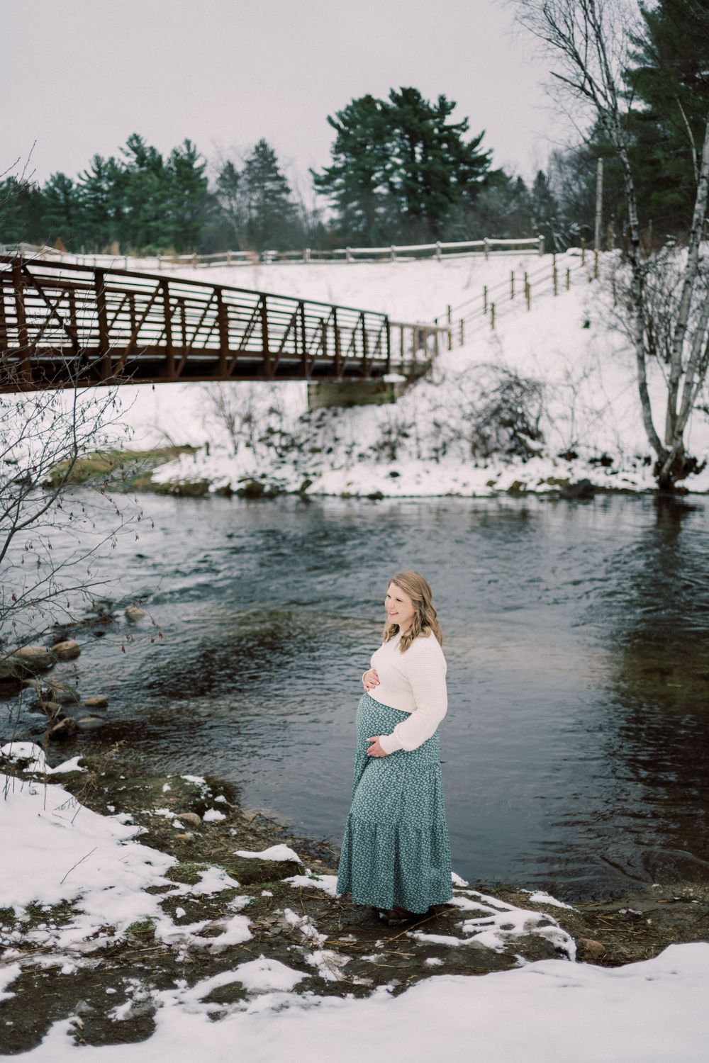 A scenic winter maternity portrait by a river with a rustic bridge in the background.