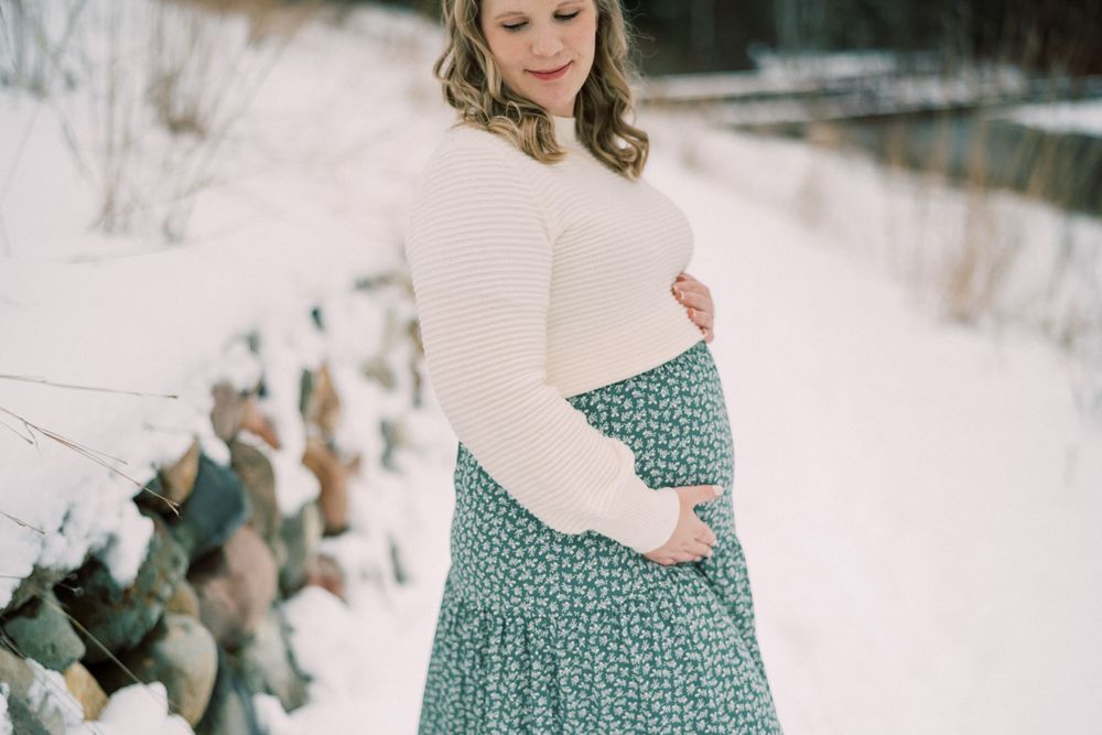 A side view maternity portrait in the snow wearing a cream sweater and patterned maxi skirt.