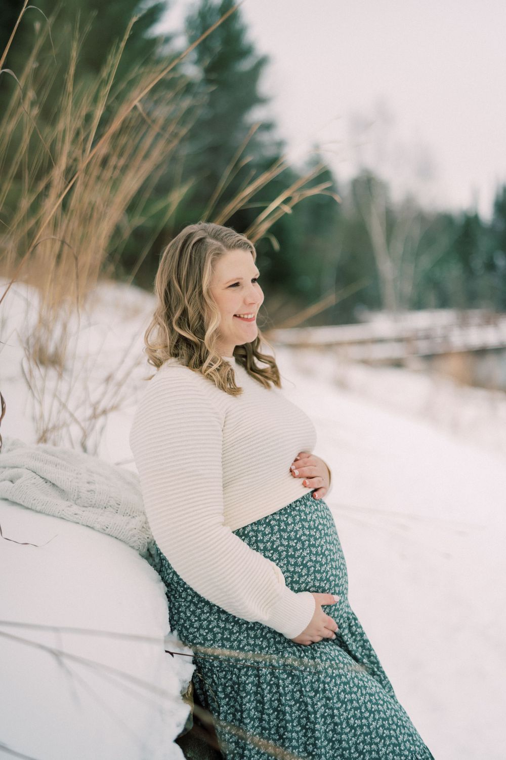 Expecting mother in white sweater and floral skirt poses in snowy winter landscape.