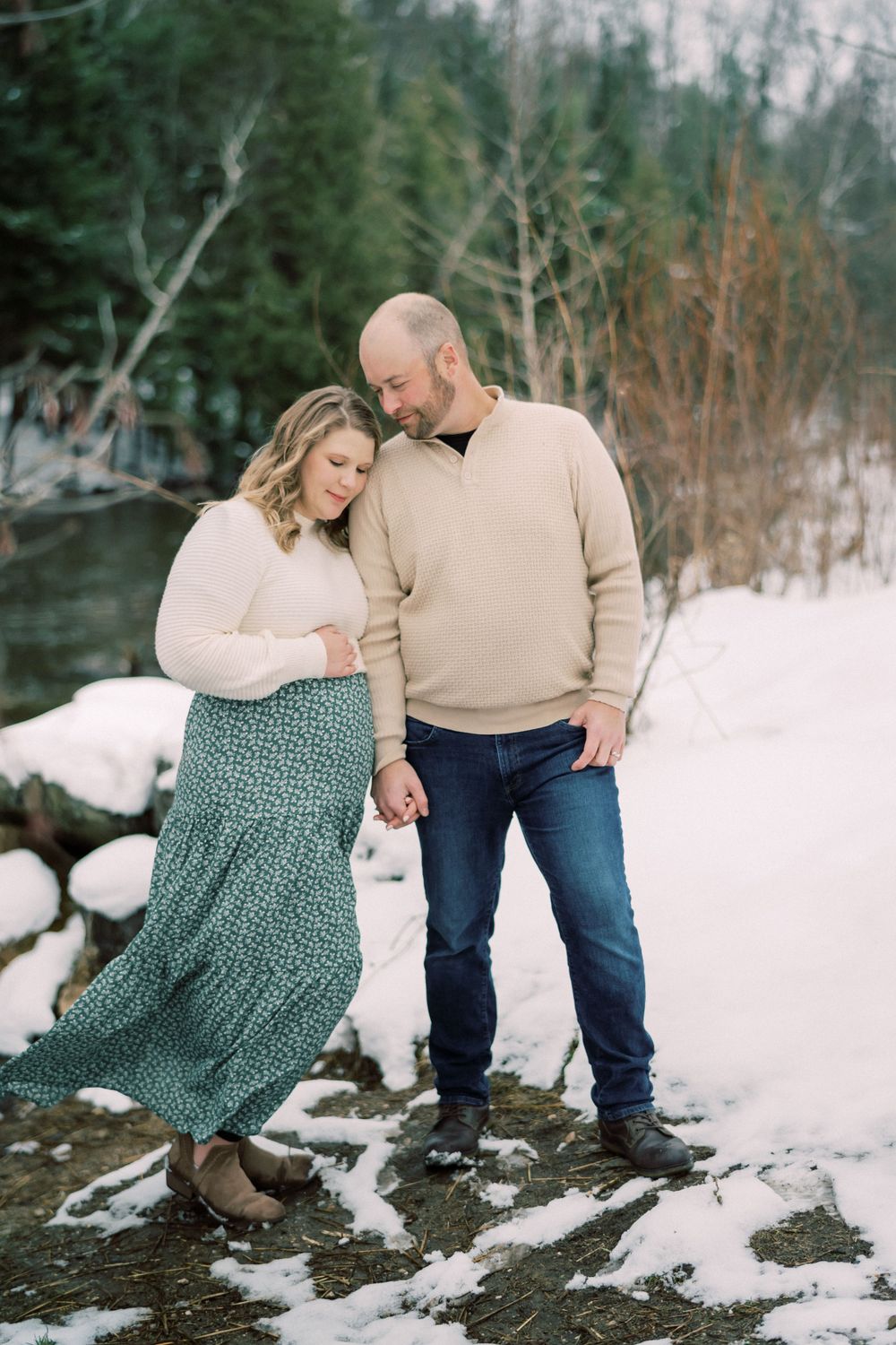 Romantic winter photo of couple in coordinating sweaters standing by snowy riverbank.