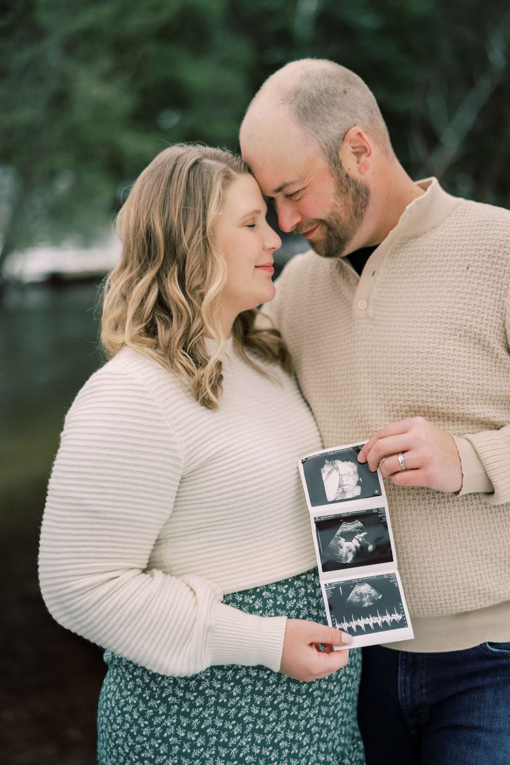 Couple sharing intimate moment while holding ultrasound photos outdoors.