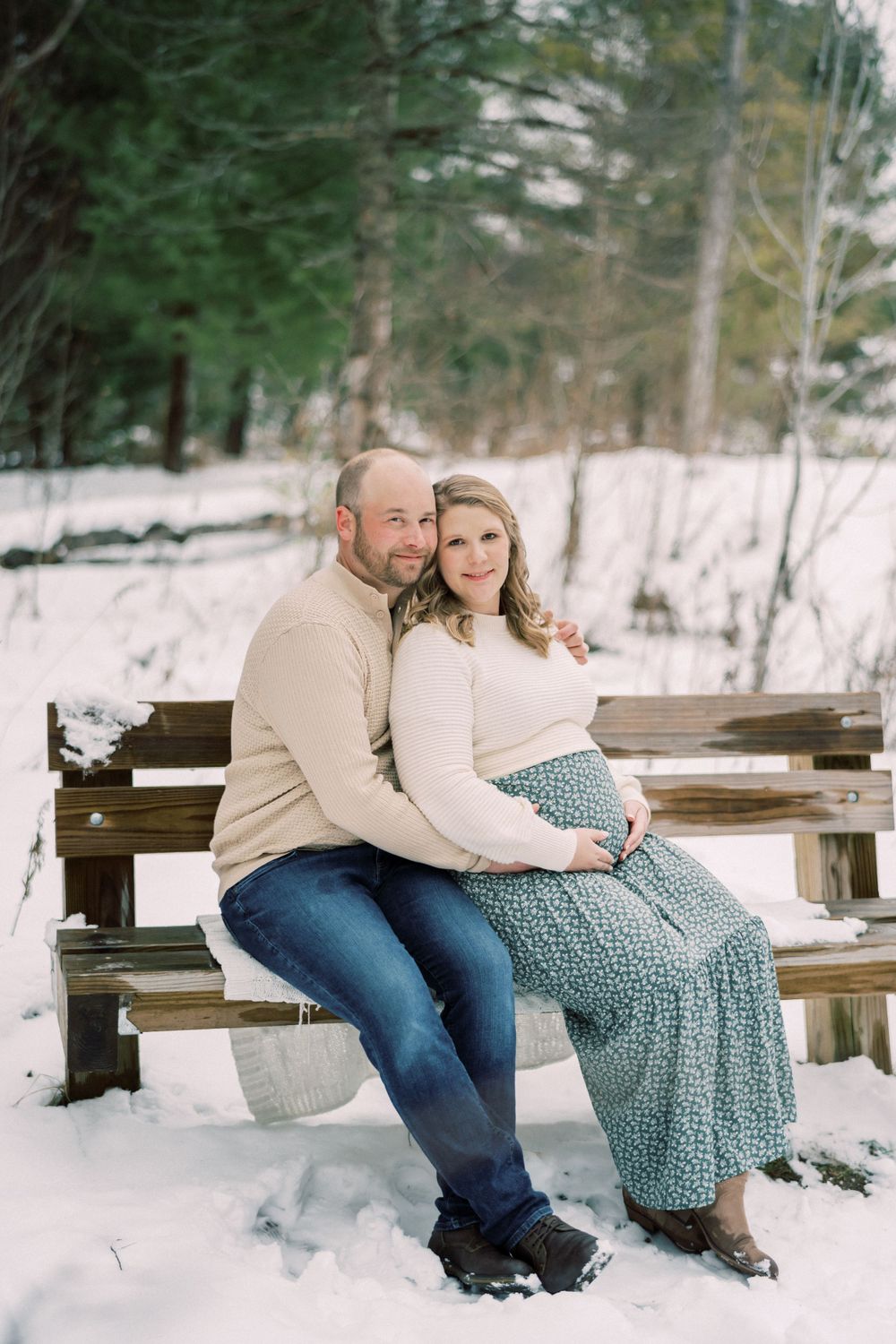 Winter maternity photo of couple sitting together on snow-covered wooden bench.