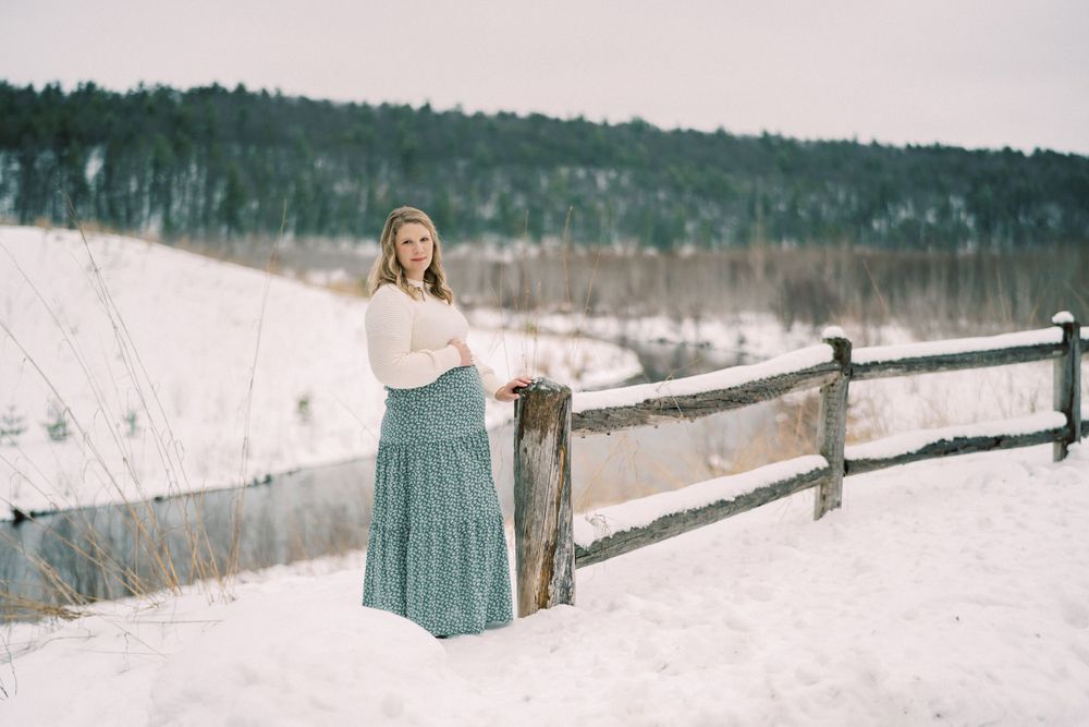 A winter maternity photo featuring a cream sweater and teal maxi skirt against a snowy landscape with wooden fence.