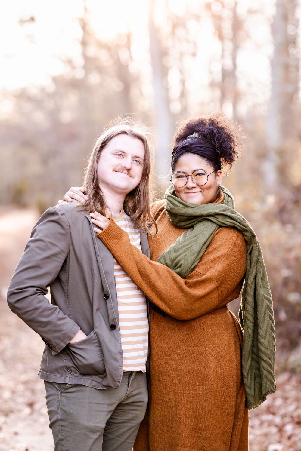 A couple in casual fall attire poses together on a wooded trail.