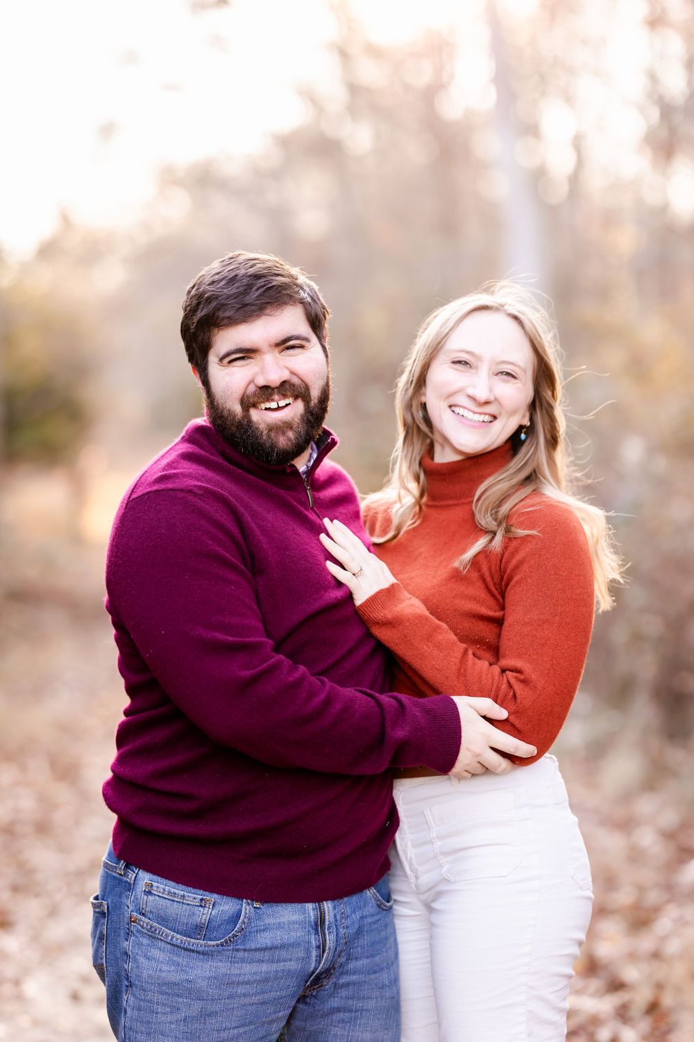 A couple in burgundy and rust sweaters embrace while posing for a fall engagement photo.