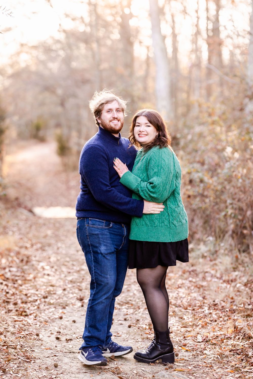 A couple in sweaters embraces on an autumn trail surrounded by fallen leaves.