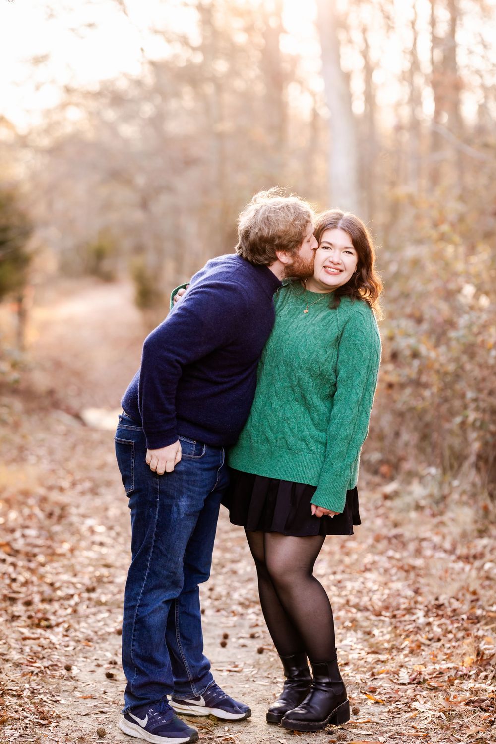 A couple shares a sweet moment on a wooded autumn trail with fall foliage in the background.