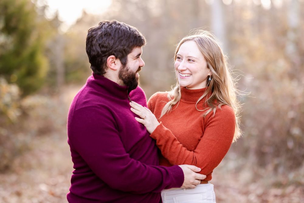 A couple in burgundy and orange sweaters share an intimate moment in an autumn forest setting.
