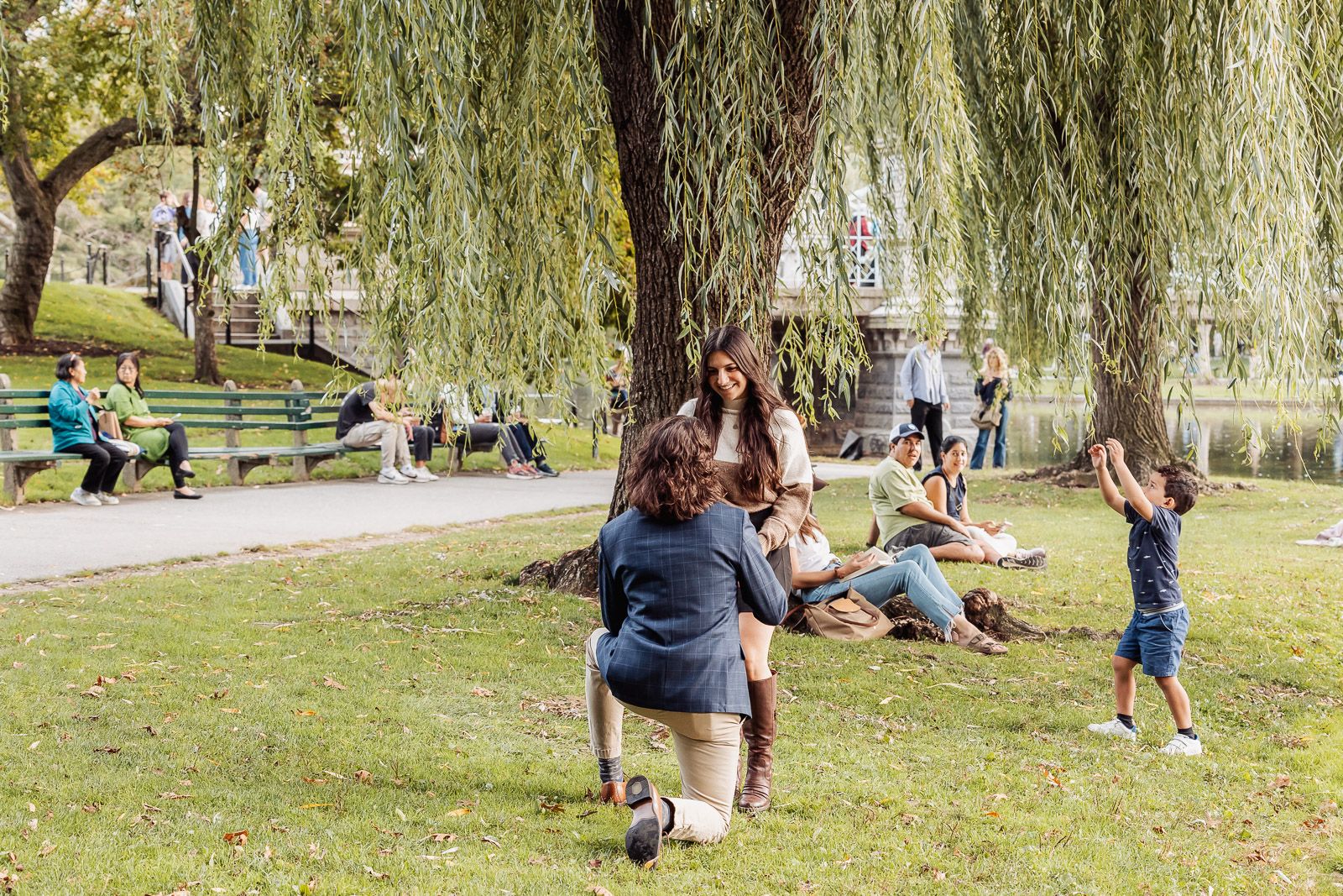 People relaxing in park under willow trees on sunny day.