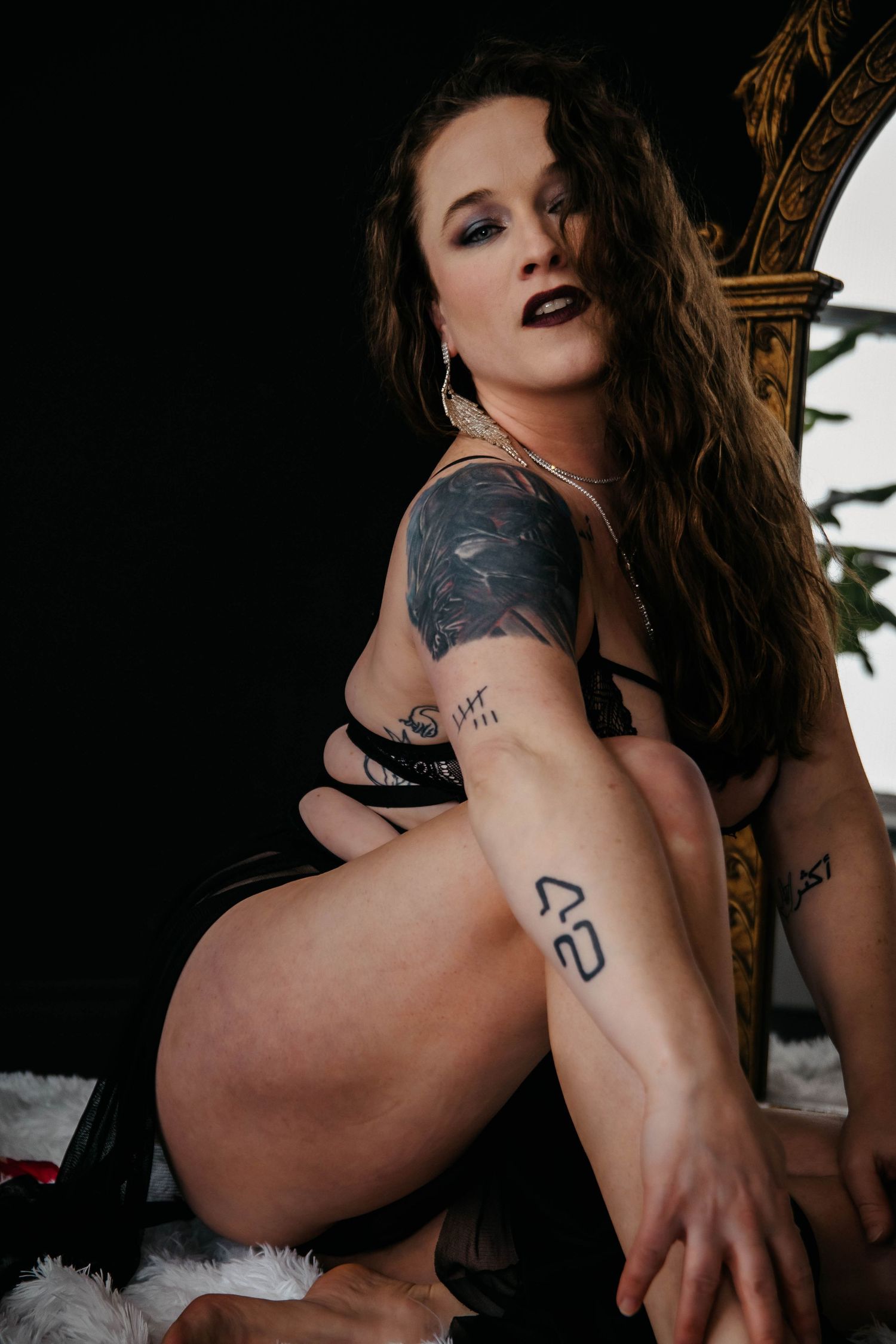 Artistic portrait of someone in black lingerie posing with visible tattoos against a dark background.