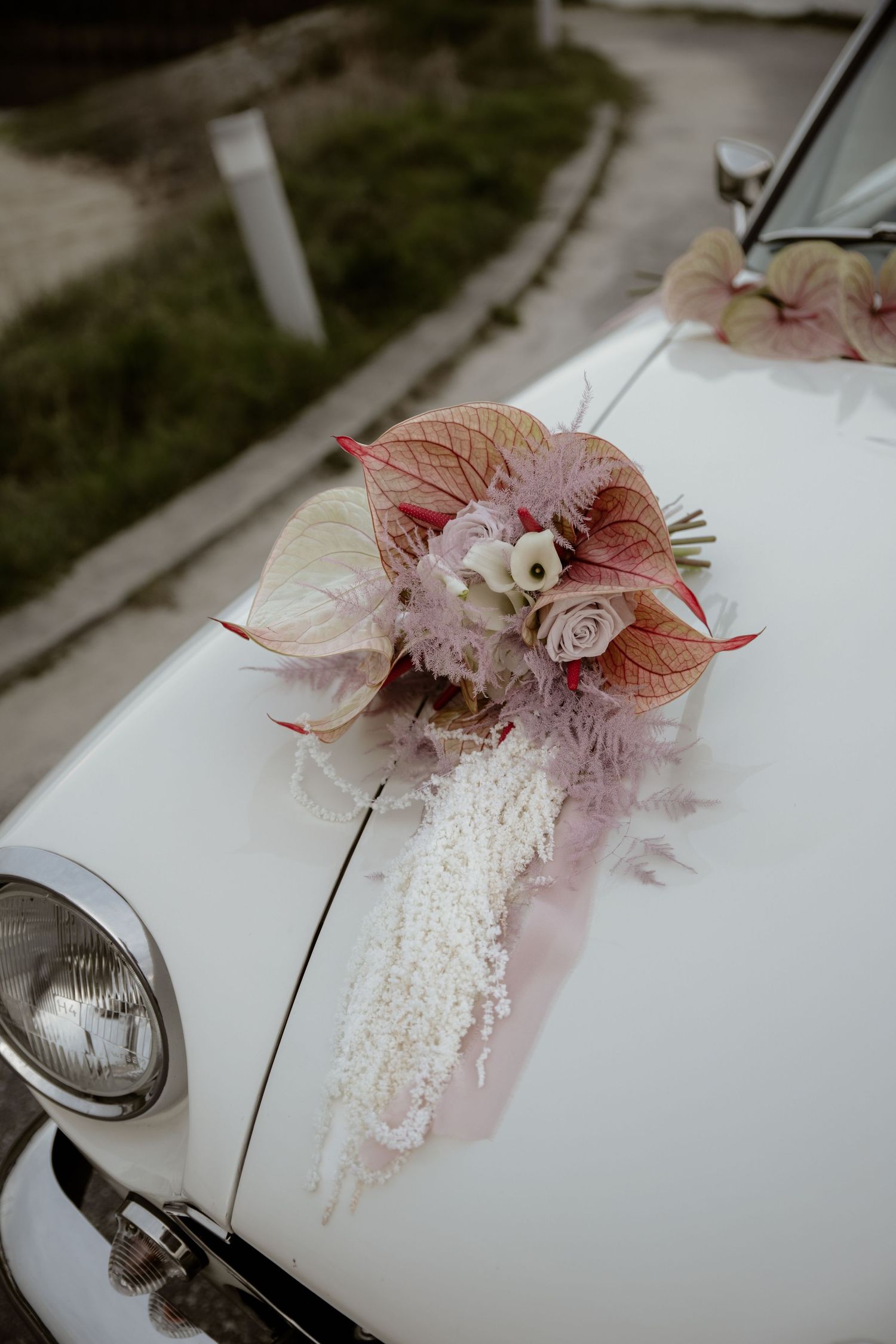 A delicate pink and white floral bouquet adorns the hood of a vintage white car.