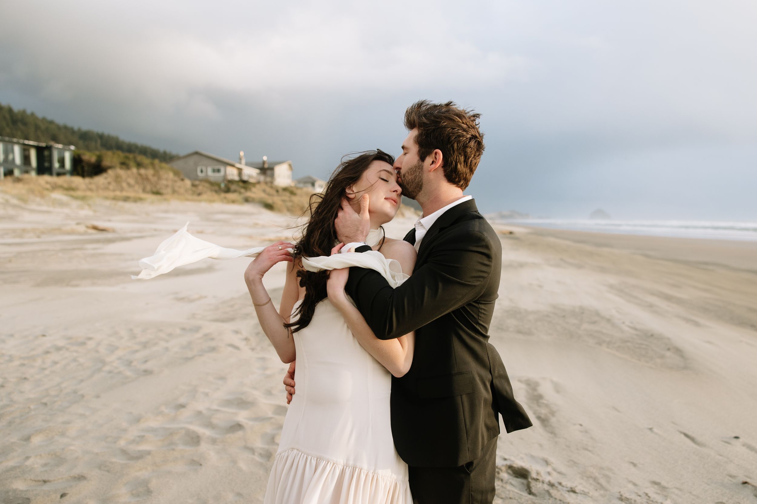 A romantic couple embraces on a cloudy beach with coastal buildings visible in the background.