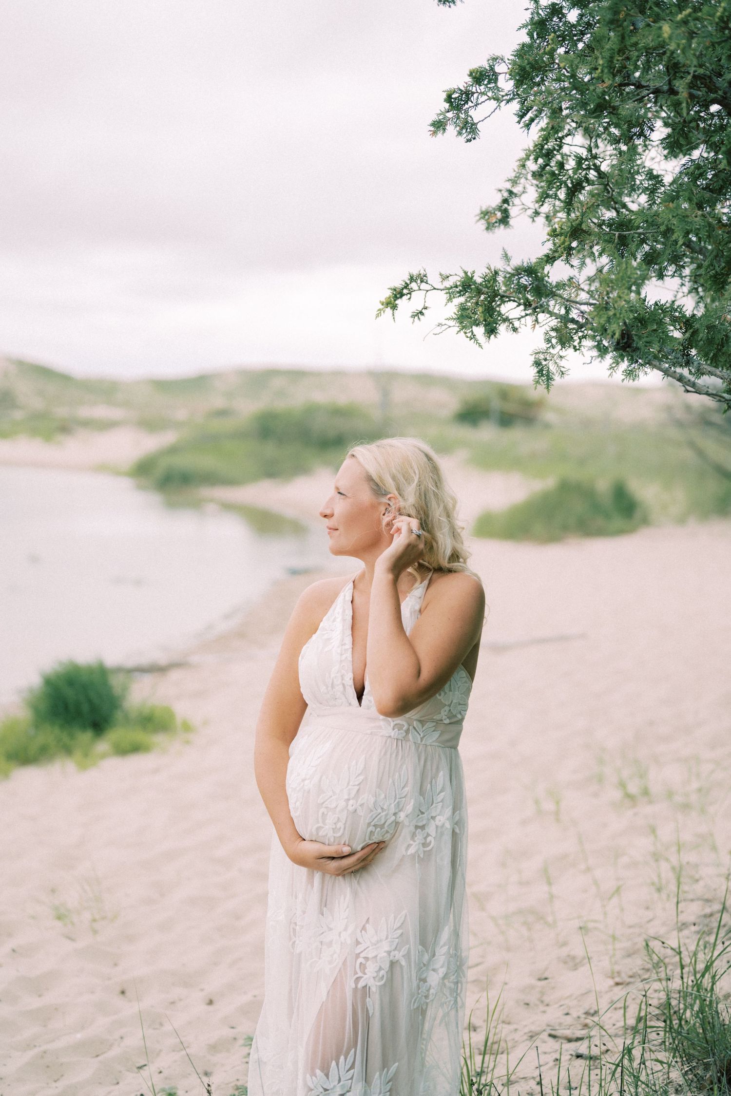 Ethereal maternity portrait of woman in white lace dress on sandy dunes.
