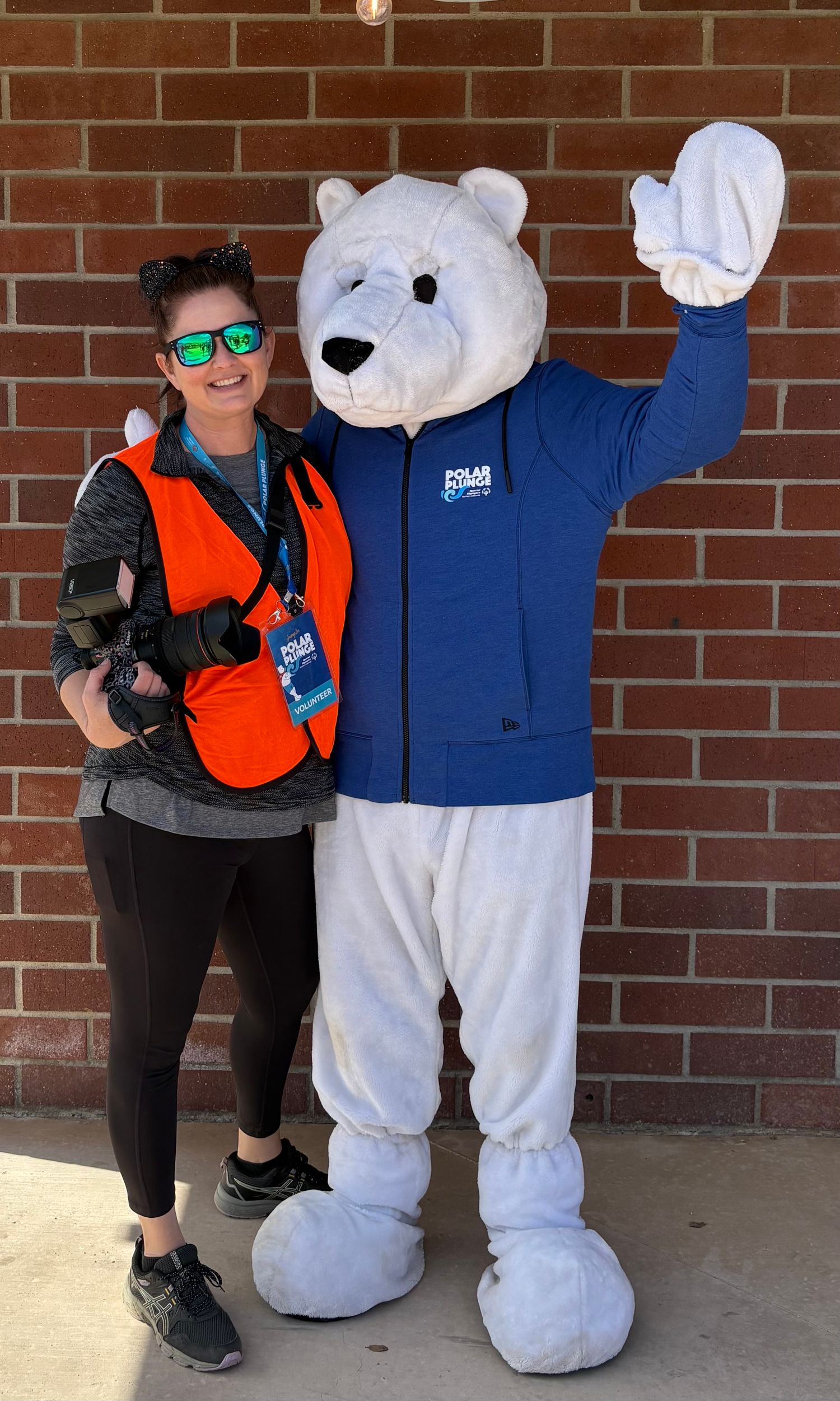 A person in orange safety vest poses with a white polar bear mascot wearing a blue jacket against a brick wall.
